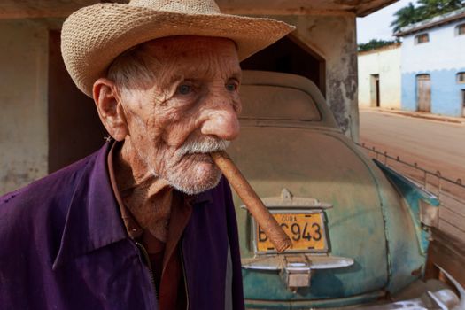 Elderly man with straw hat and cigar in front of vintage car on a Cuban street.