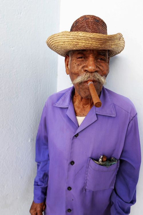 Free Senior man with mustache wearing hat and purple shirt, smoking cigar against white wall. Stock Photo