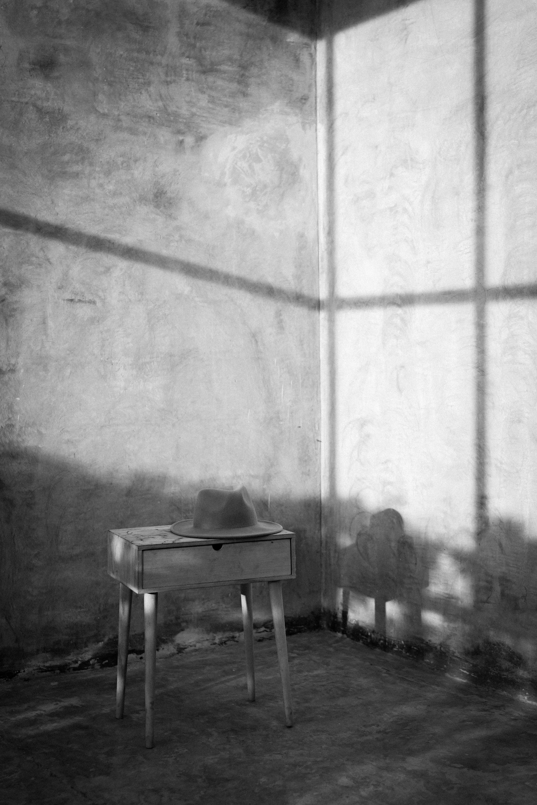 Free A minimalist black and white photo of a bedside table with a hat casting shadows on a wall. Stock Photo