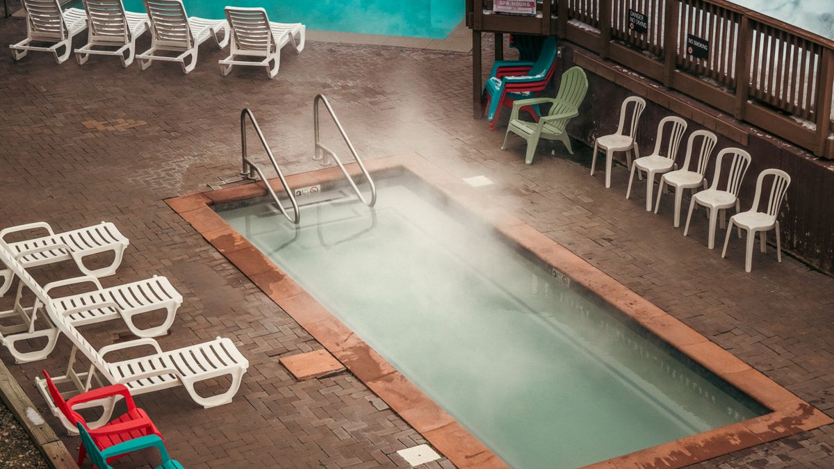 Aerial view of a steaming swimming pool surrounded by sunbeds and colorful chairs on a paved area.