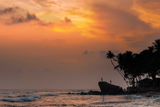 Dramatic sunset silhouette of two figures on a rocky shore at Unawatuna Beach, Sri Lanka.