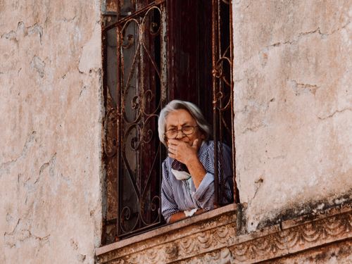 Free Elderly woman with gray hair and glasses leaning out a rustic window, contemplating. Stock Photo