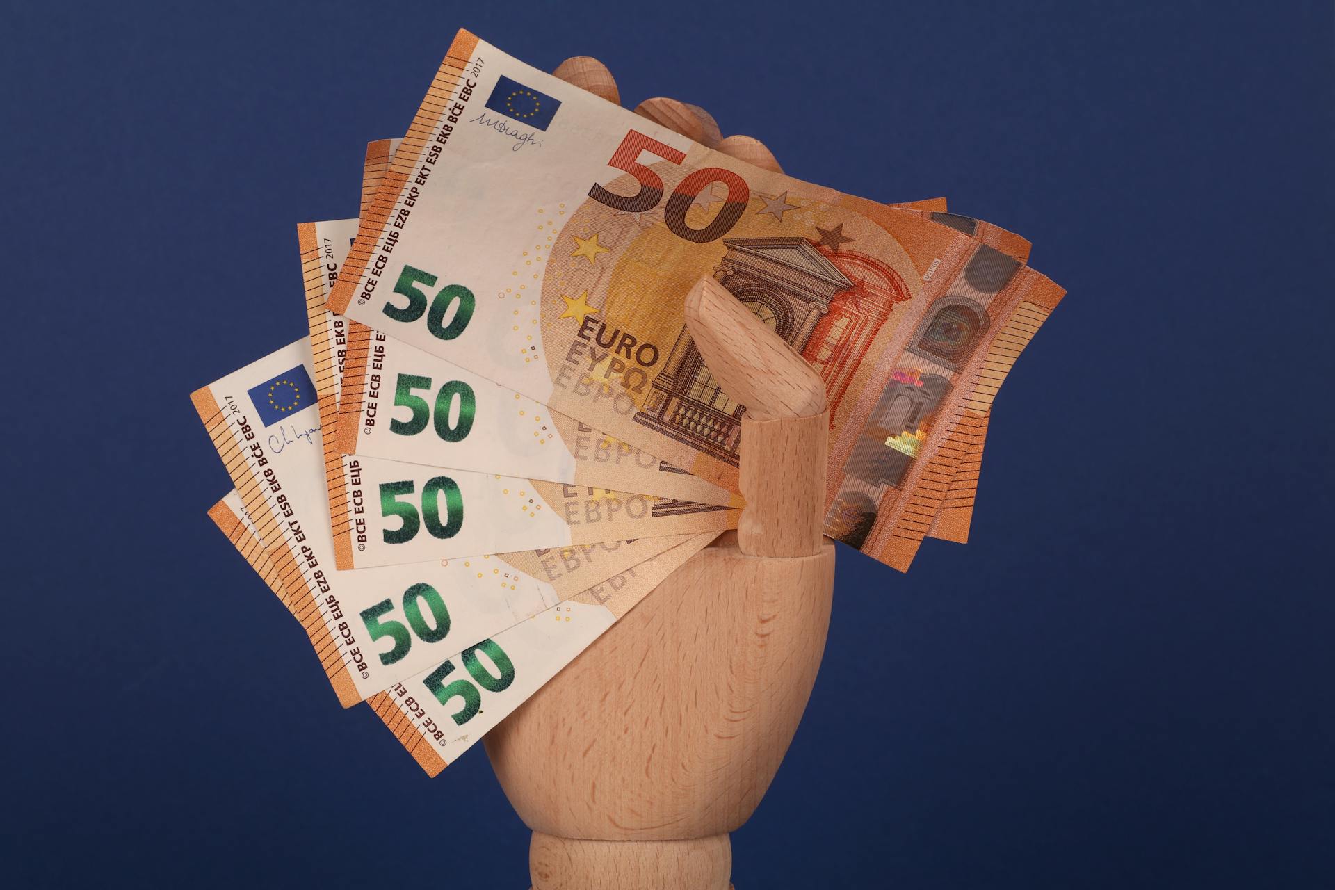 A wooden mannequin hand grasping fifty euro banknotes with a blue backdrop.