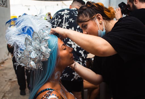 A makeup artist prepares a carnival performer with detailed face makeup and feathers.