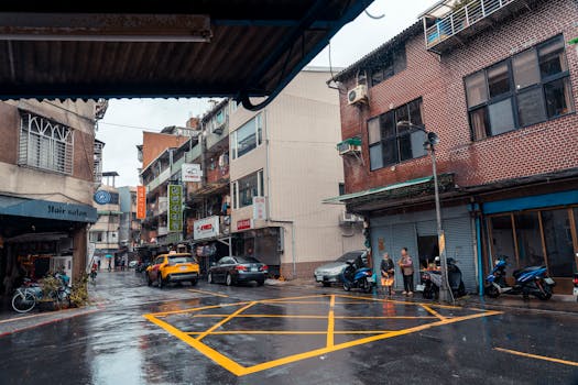 A wet intersection in an urban area featuring cars and pedestrians under a cloudy sky.