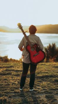 Person with a guitar watching a serene sunset over a lake. Perfect for music and nature themes.