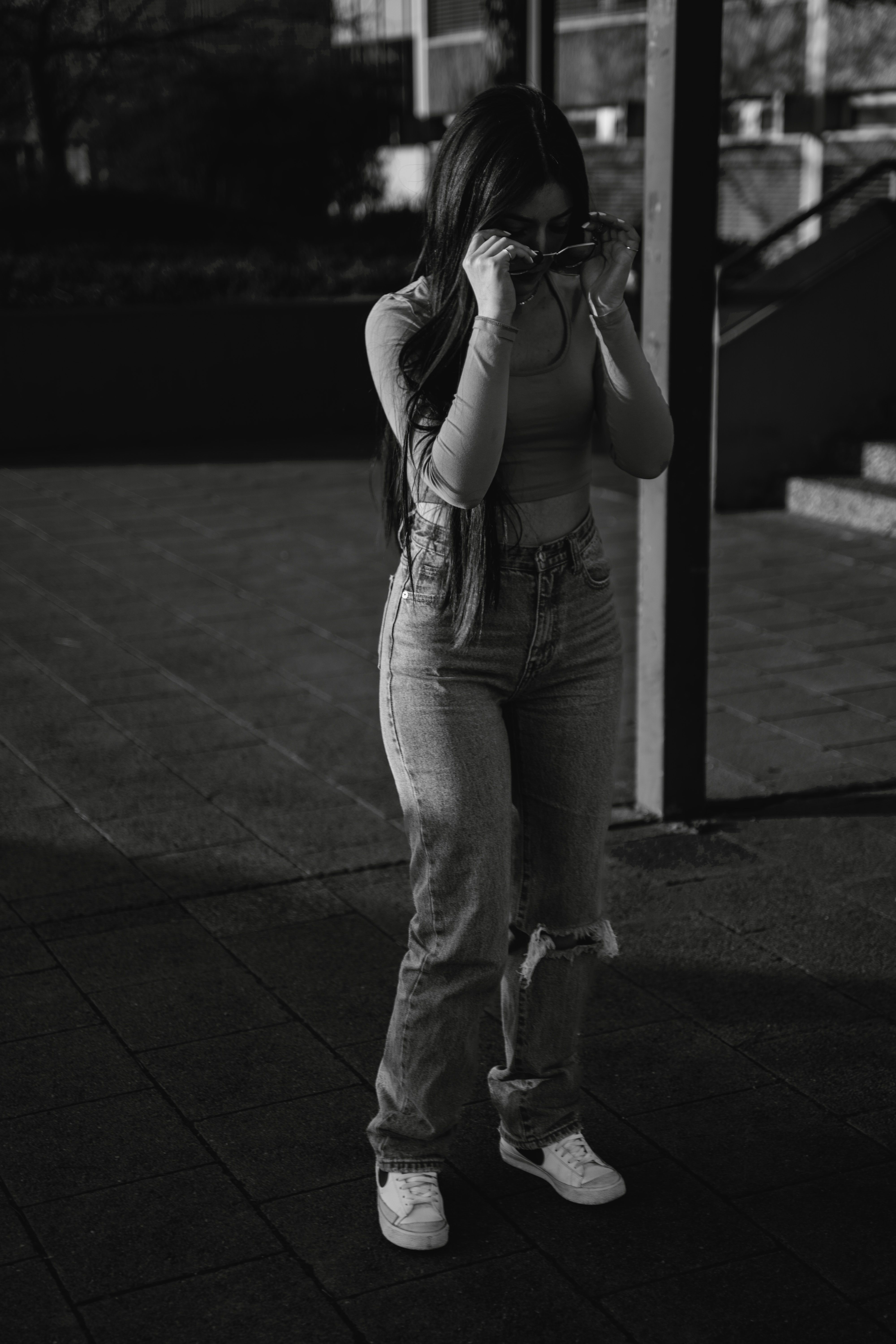 Black and white photo of a woman in casual wear adjusting sunglasses outdoors on a pavement.
