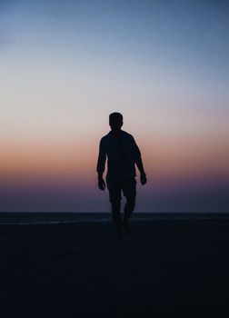 A solitary man walks on a beach as twilight sets, creating a striking silhouette against a colorful sky.