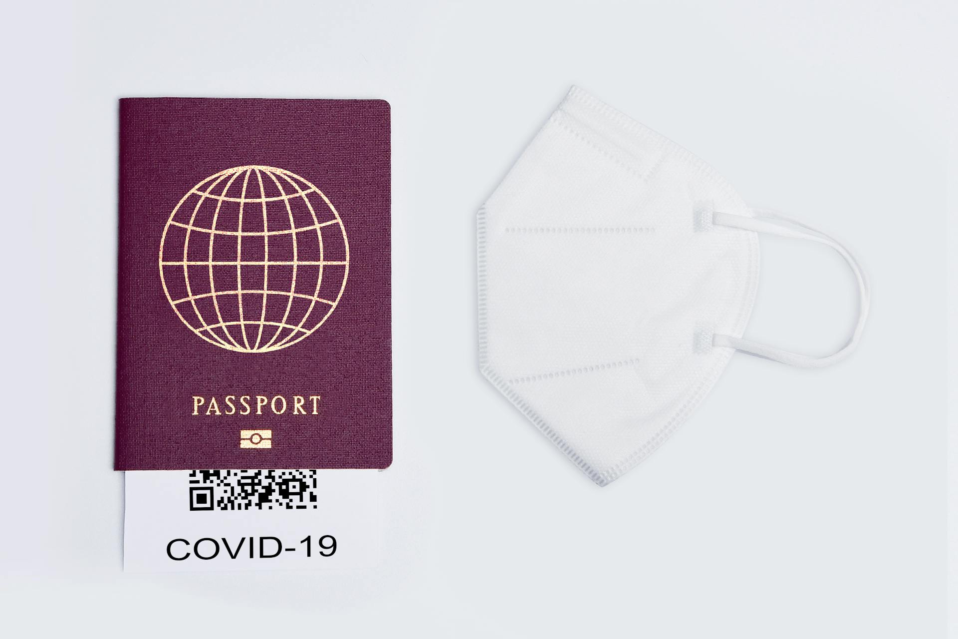 Flat lay of a travel passport, face mask, and COVID-19 QR code on a white surface, symbolizing pandemic travel.