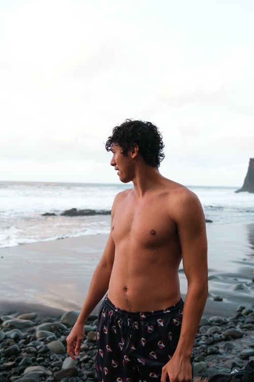Free A topless man stands on a rocky beach gazing at the ocean waves. Stock Photo