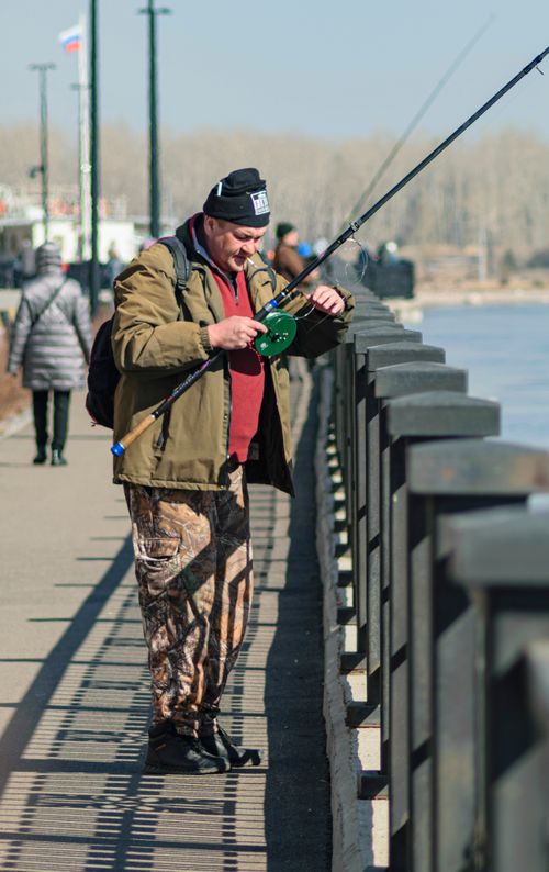 Free A man wearing winter clothing fishes from a riverbank with a fishing rod on a sunny day. Stock Photo