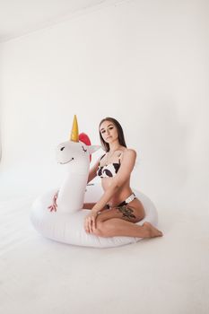 Woman sitting on a unicorn pool float, wearing a swimsuit in a bright studio setting.