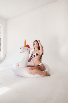 Young woman in bikini posing with inflatable unicorn indoors, showcasing tattoo art.