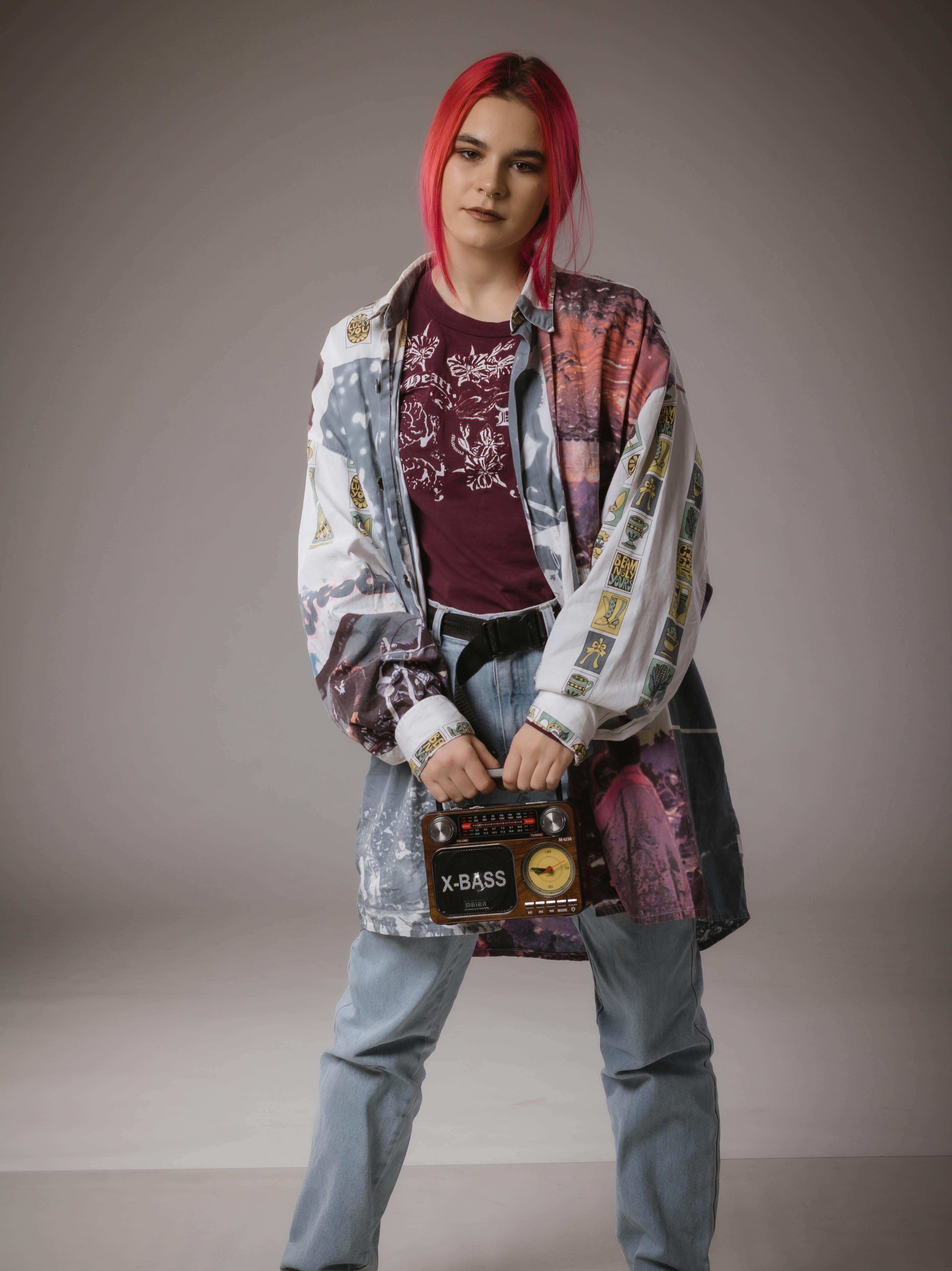 Stylish woman with red hair holding a retro radio, wearing denim jeans and a printed jacket in a studio setting.