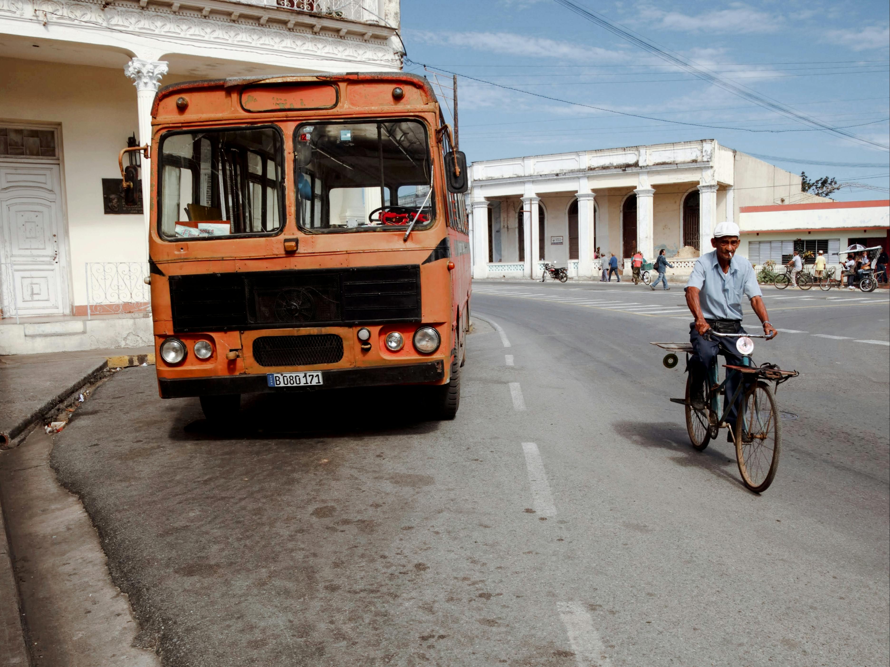 Free A man rides a bicycle past a parked bus on a city street, showcasing urban life. Stock Photo