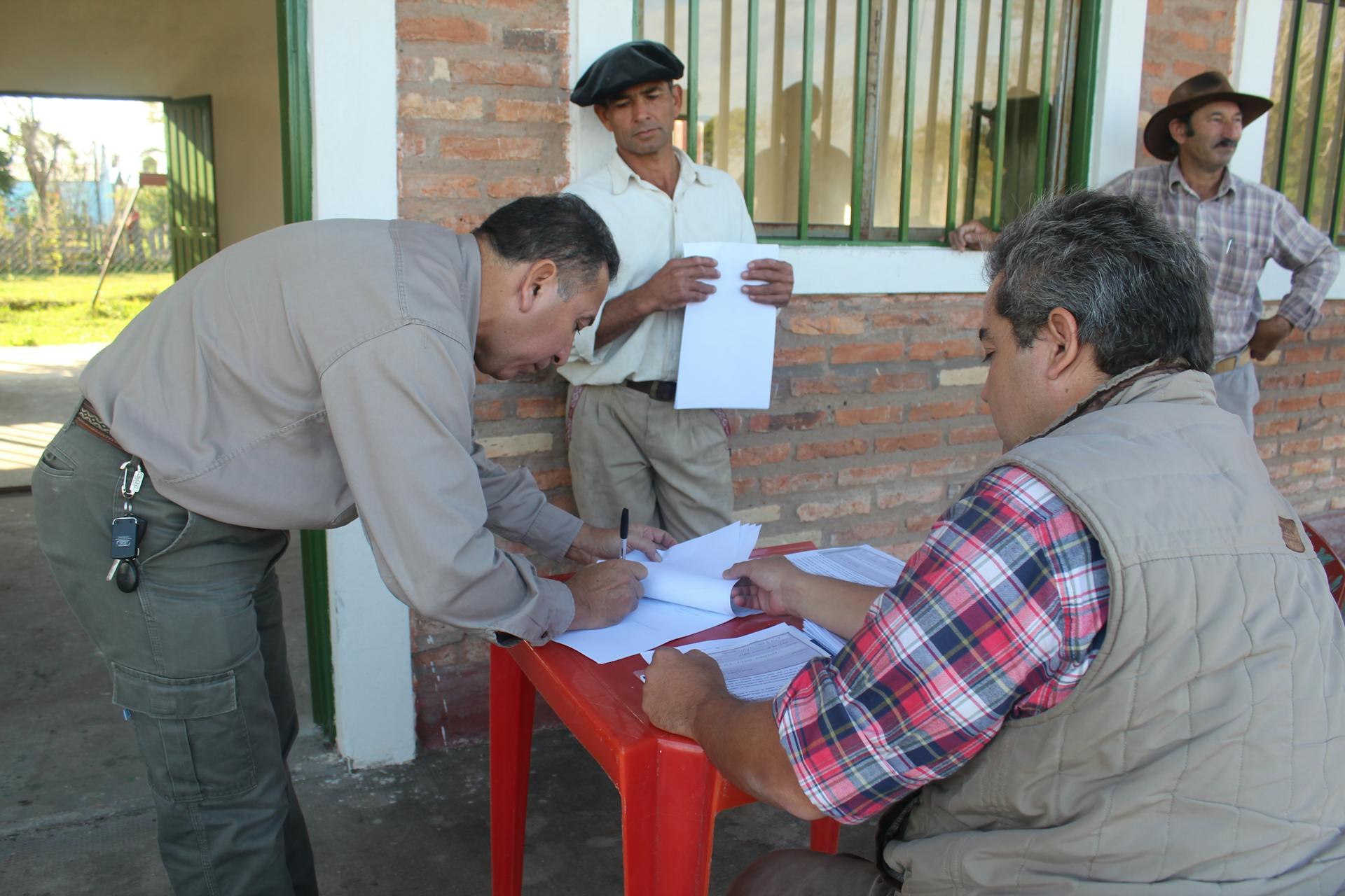 Men conducting paperwork outdoors, signing documents at a table near a brick building.