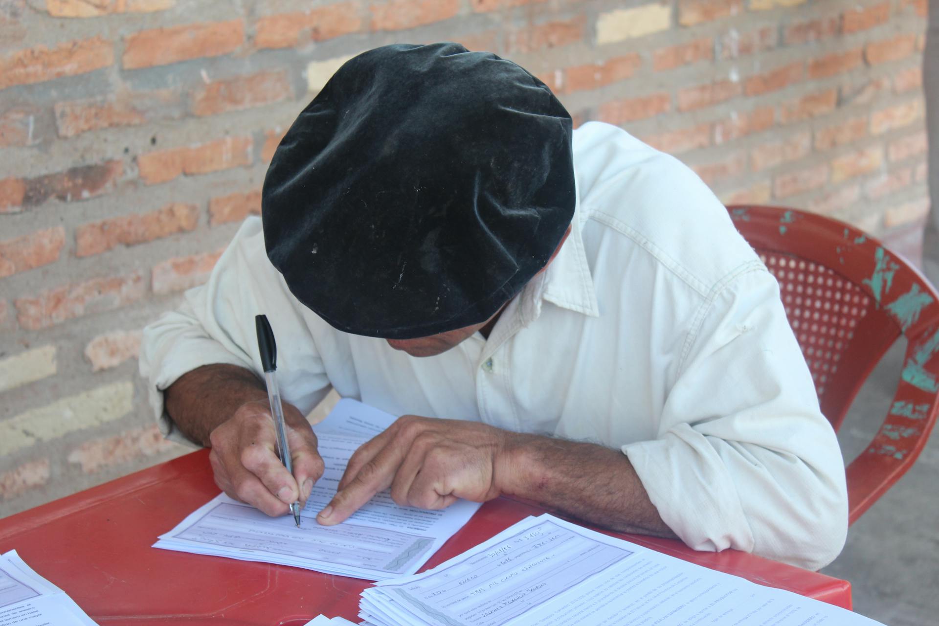 Male adult writing on papers wearing a flat cap, indoors with a brick wall backdrop.