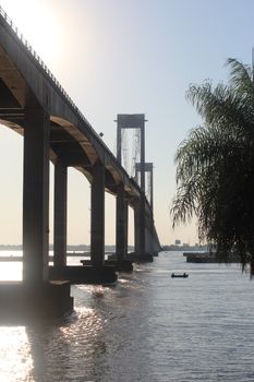 A scenic view of a suspension bridge over a river with a clear sky and palm tree at sunrise.