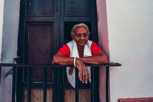 Free Elderly woman in red leaning on a balcony railing, looking serene. Stock Photo