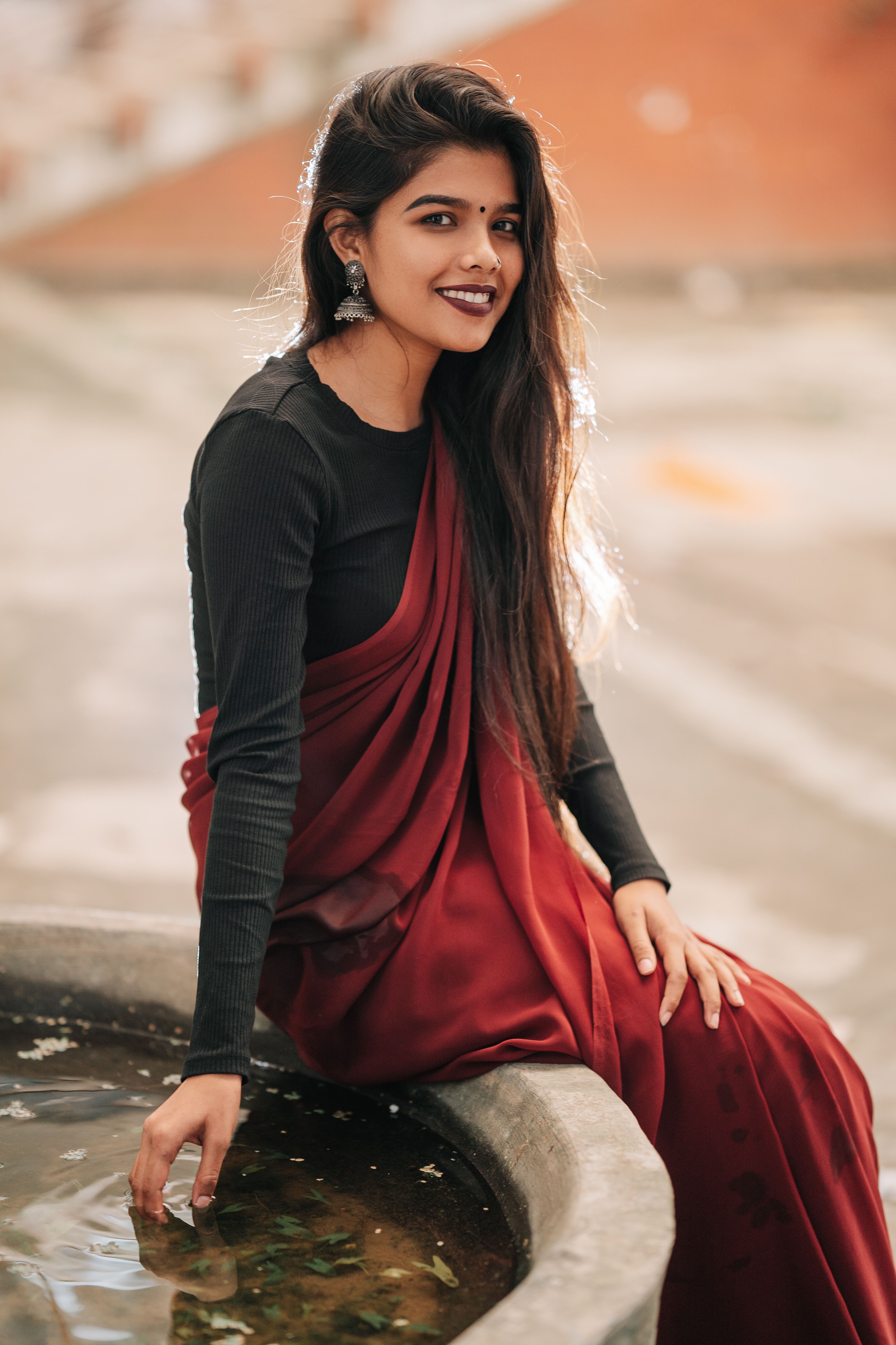 A woman wearing a red saree smiles while seated near a water feature outdoors.