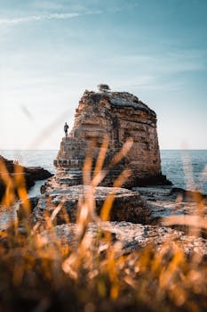 Lone person standing on a towering coastal rock with sea view, under clear skies.