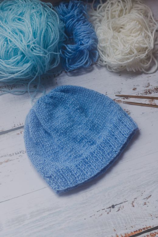 A blue hand-knitted beanie surrounded by skeins of yarn on a rustic wooden table