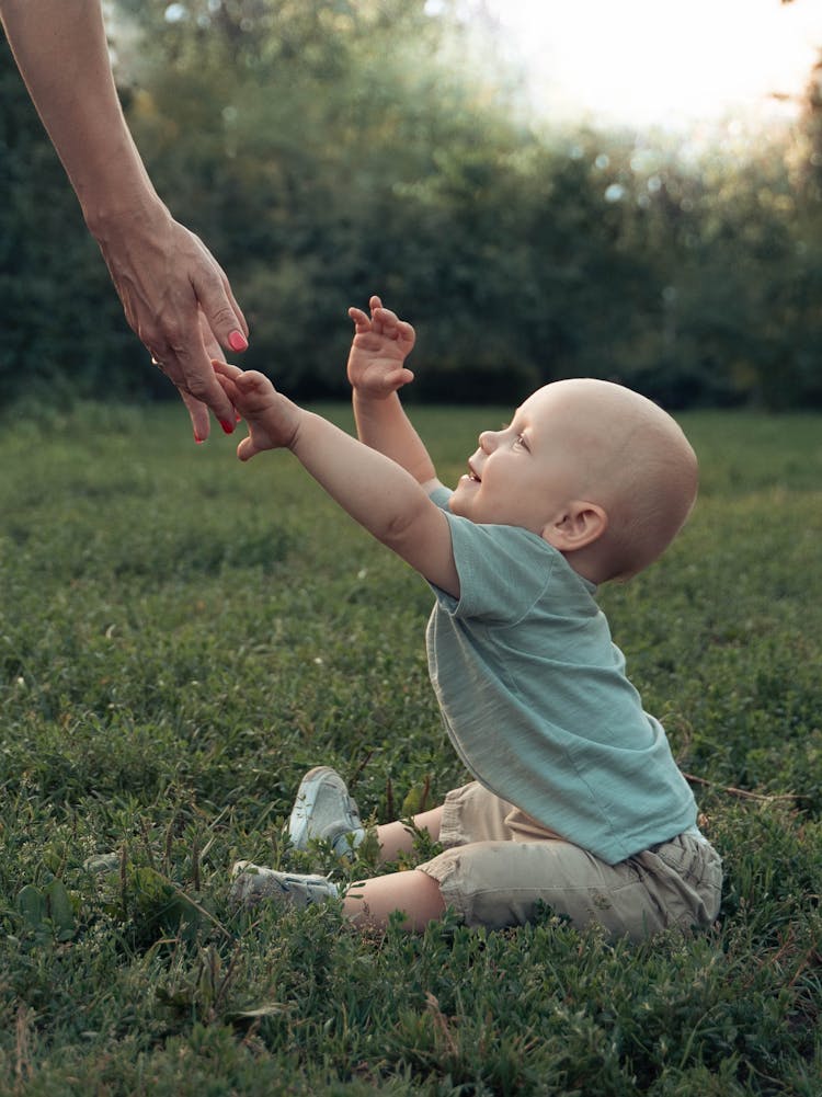 Baby Boy Sitting On Grass And Reaching For His Mothers Hand