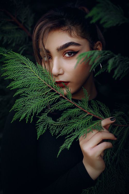 Free Elegant portrait of a woman with bold makeup, partially hidden behind vibrant green leaves. Stock Photo