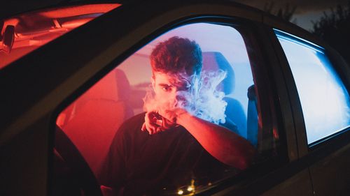 Free Moody image of a young man vaping inside a car with vibrant red and blue lighting. Stock Photo