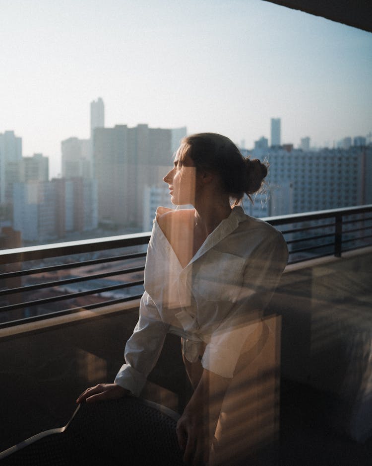 Photo Of Woman Standing On The Balcony Looking At The City From Behind The Window