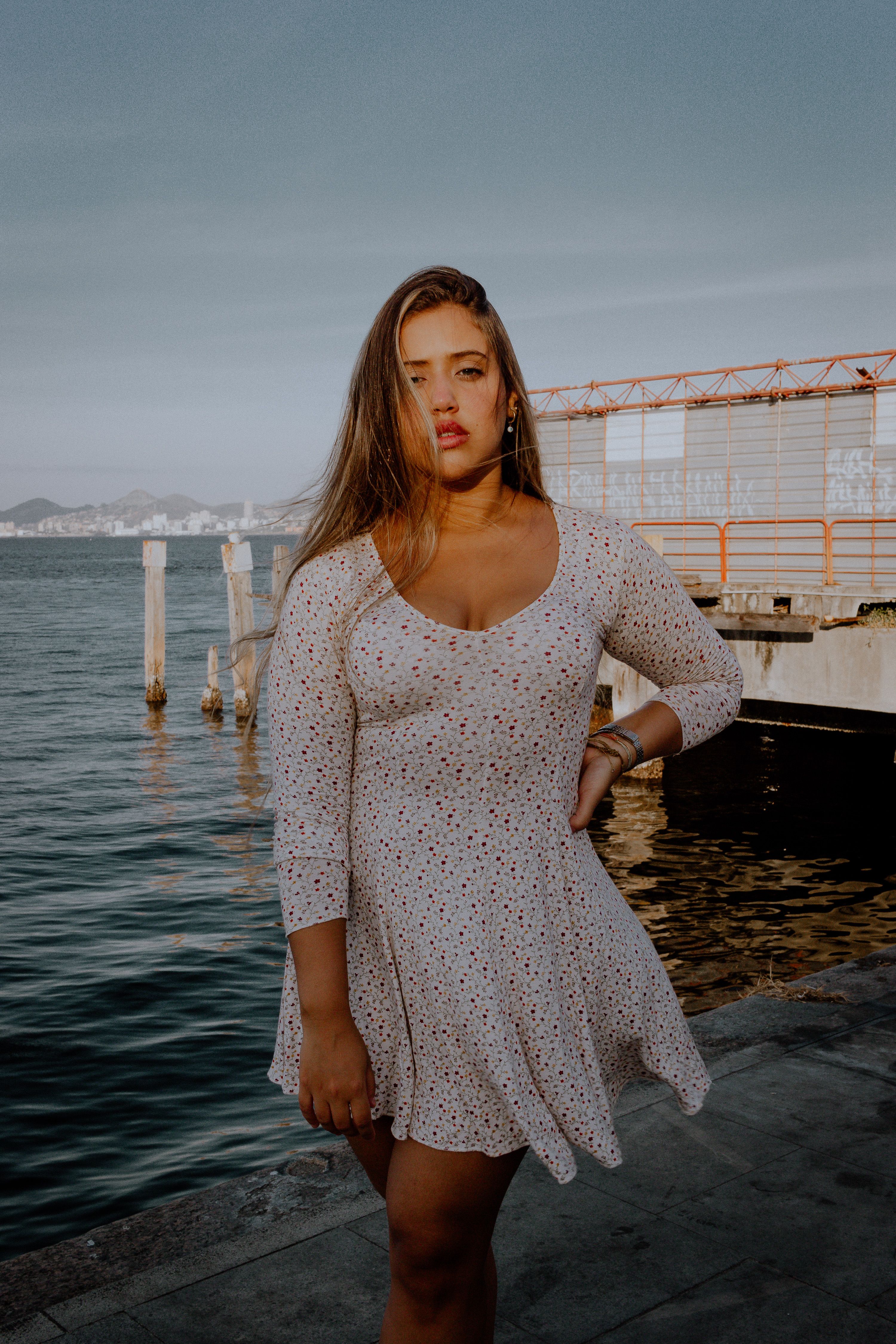 A woman in a floral dress poses confidently by the waterfront in daylight.