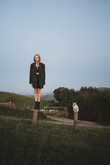 A woman in an oversized coat stands on a post beside a cat in a pastoral landscape.