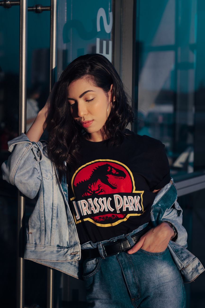 Stylish woman in graphic tee and denim jacket posing against a glass door, bathed in sunlight.