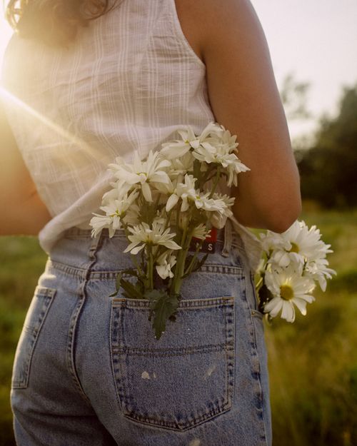 Free A woman stands outdoors with flowers in her jeans pocket, bathed in warm sunset light. Stock Photo