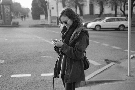 Woman in sunglasses smiling at phone on a sidewalk, captured in monochrome.