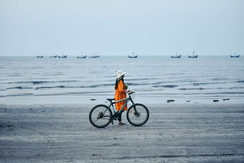 Free A woman in an orange dress walks her bicycle along a tranquil beach with boats in the distance. Stock Photo