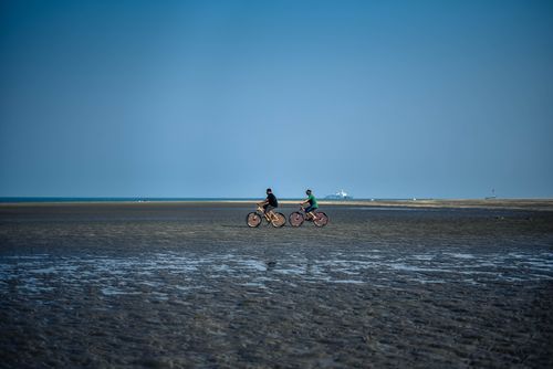 Free Two men cycling on a beach during low tide under a clear blue sky. Stock Photo