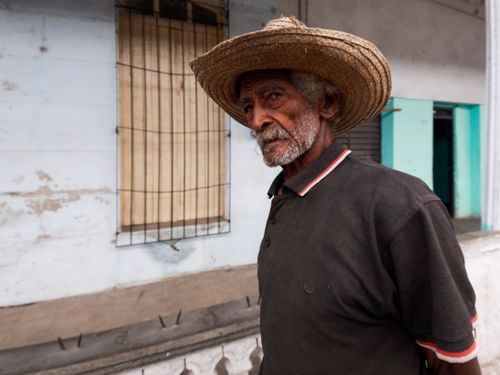 Free Portrait of an elderly man with a straw hat standing beside a rural building, showcasing a rustic lifestyle. Stock Photo
