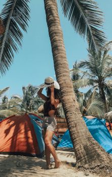 A woman in a hat leans against a palm tree near colorful tents on a sunny beach in the Philippines.