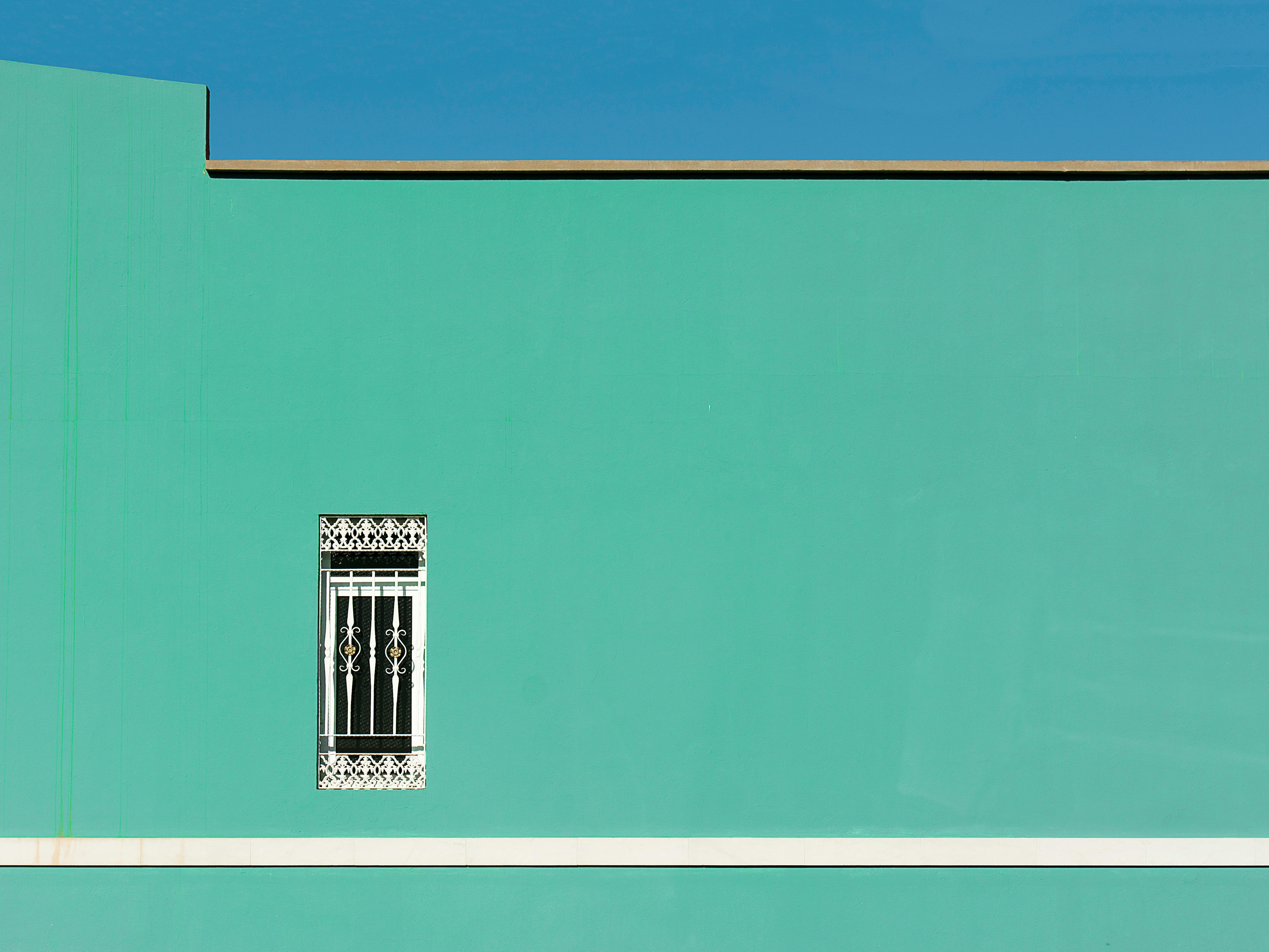 A vibrant minimalist green wall featuring an ornate window against a clear blue sky.