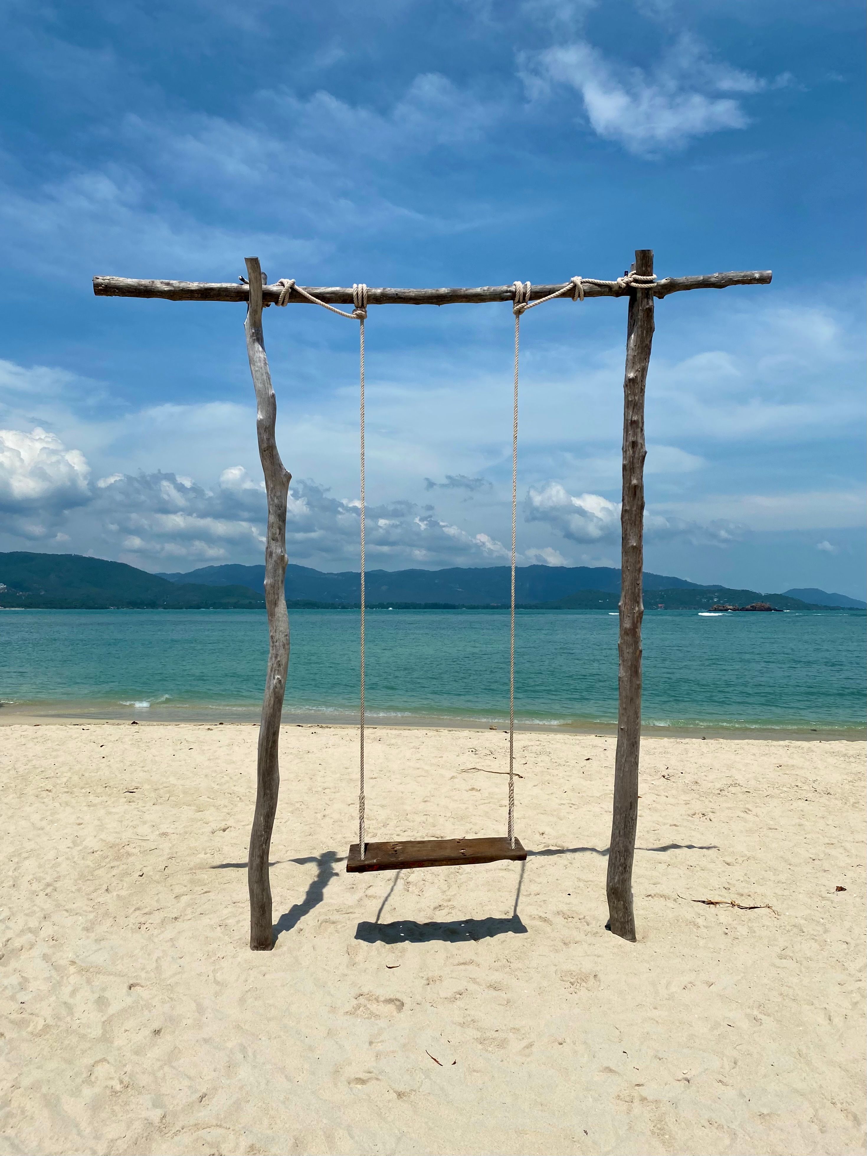 Wooden swing on a sandy beach with clear ocean and blue sky, perfect for relaxation.