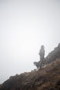 Man in Blue Jacket Standing on Brown Rock Mountain With Black Dog
