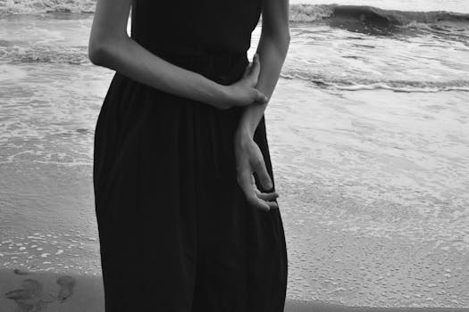 A woman in a black dress stands pensively by the ocean shore in monochrome.