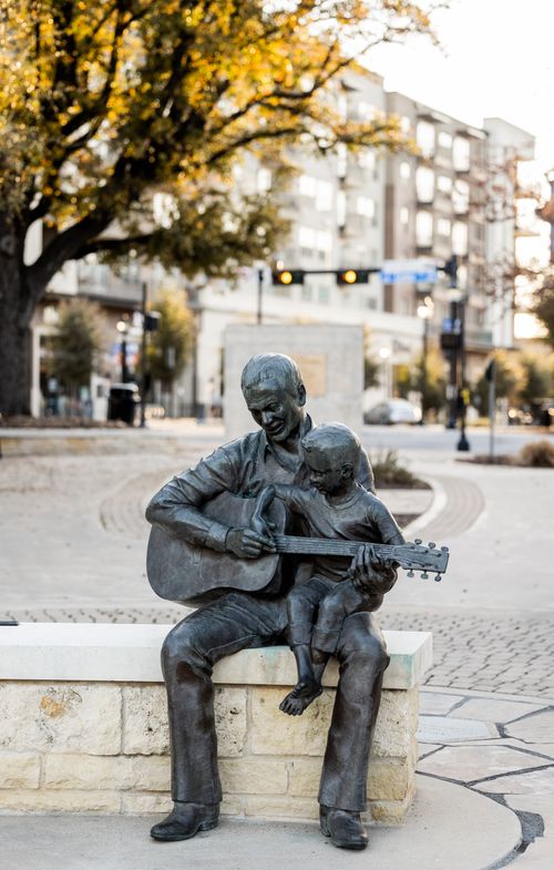 Free Bronze statue depicting a man and child sharing a musical moment with a guitar, set outdoors. Stock Photo