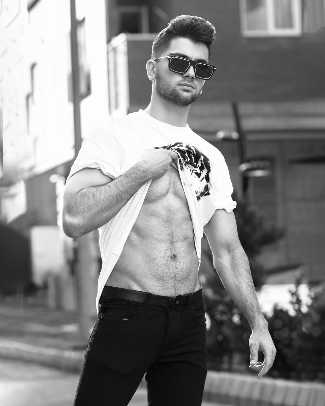 A fashionable man showing off his toned abs in an urban setting, wearing sunglasses.