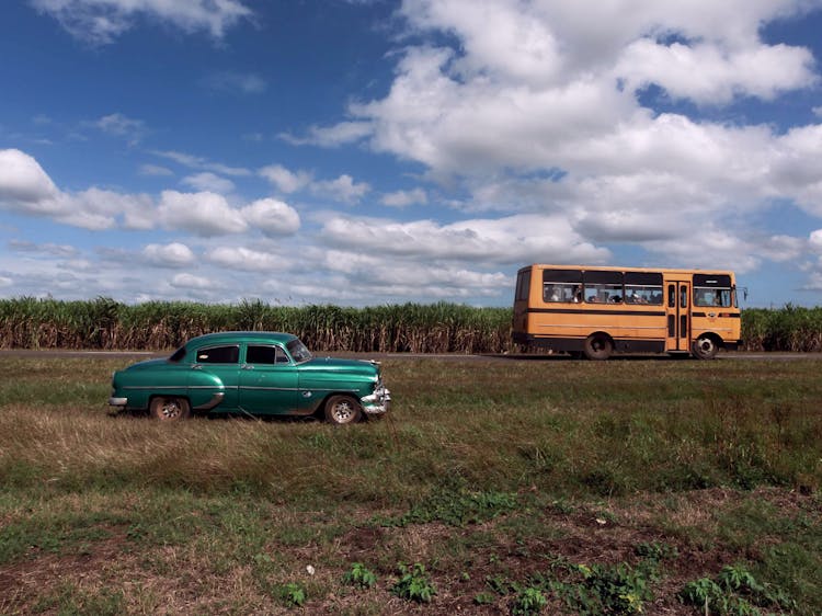 People Riding On A Yellow Bus Near A Green Vintage Car Parked On A Grass Field