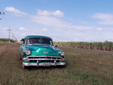 Classic green car parked on a country road surrounded by fields and blue sky.