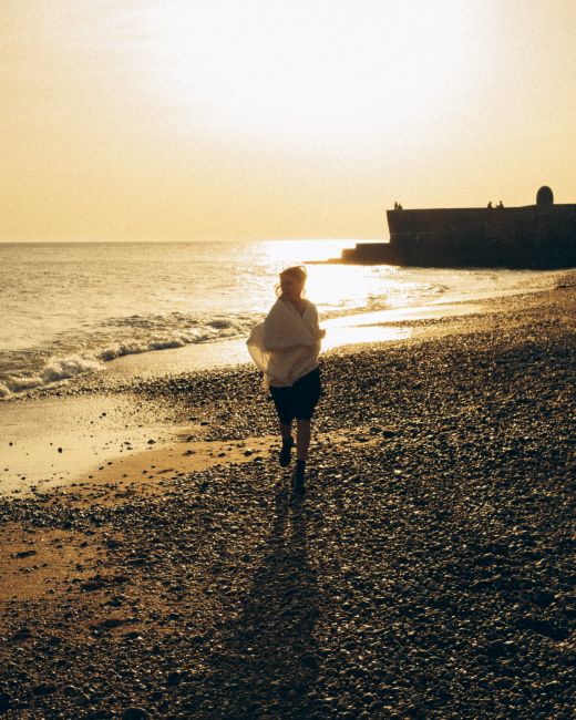 A woman runs along a rocky beach during a vibrant sunset, casting long shadows.