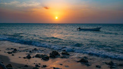Free Peaceful sunset at a rocky beach with waves lapping the shore and a lone boat on the horizon. Stock Photo