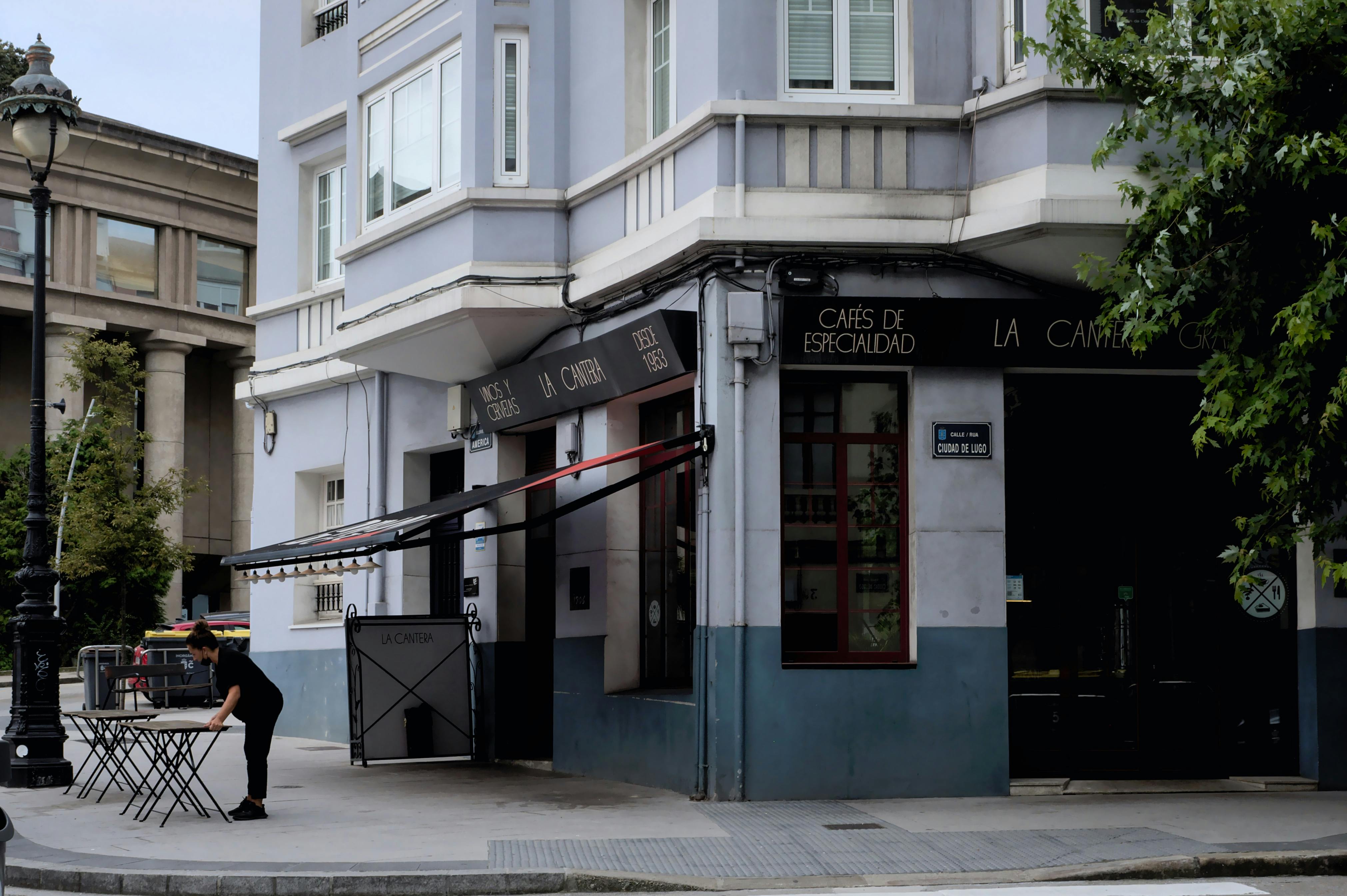 Free Street view of a café in A Coruña, showcasing urban life with a waitress setting tables. Stock Photo